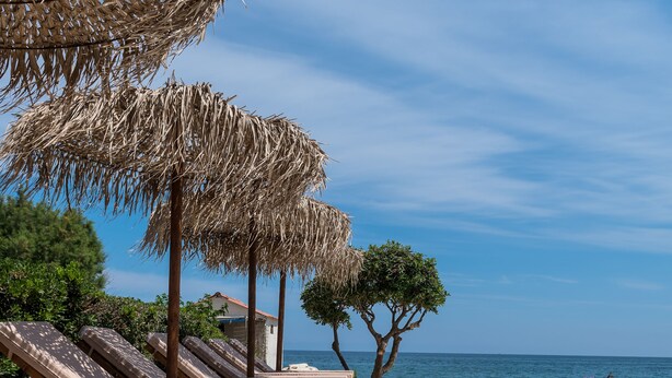 A beachside lounge area with sun loungers and thatched umbrellas, a clear blue sky, and a view of the sea. - Musela Studios (Rethymno)