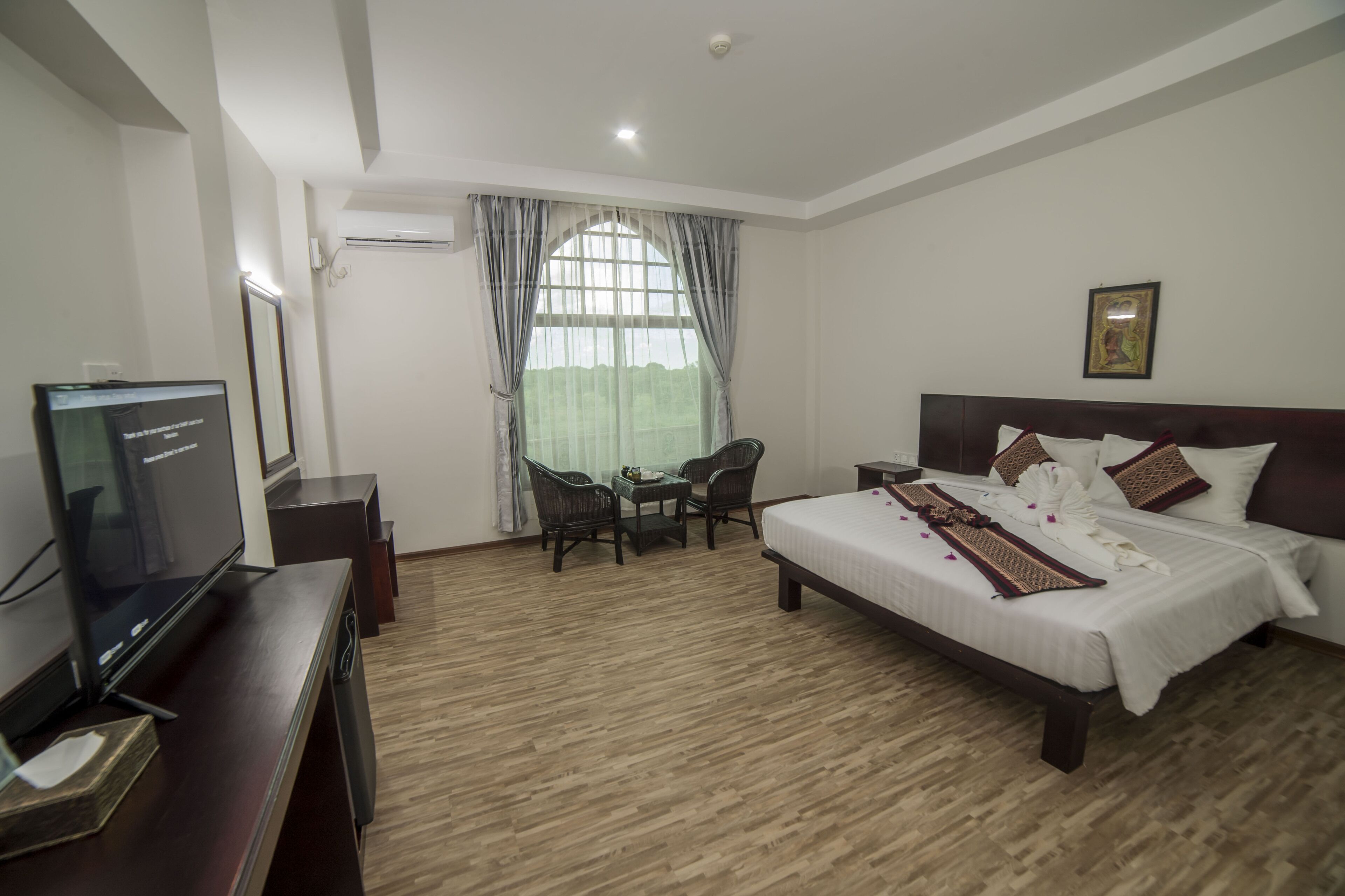 Habitación de hotel con cama, escritorio con televisor, ventana con cortinas e imagen enmarcada en la pared.
