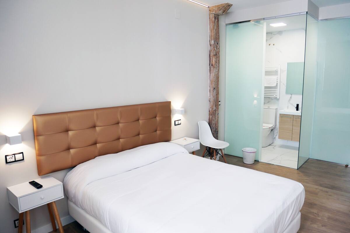 A modern bedroom with a large bed, bedside tables, a chair, and a bathroom visible through a glass door.