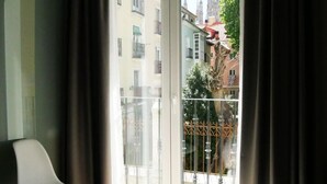 Ett hotellrum med en stor säng, en vit stol och en balkong med utsikt  över byggnader och träd.