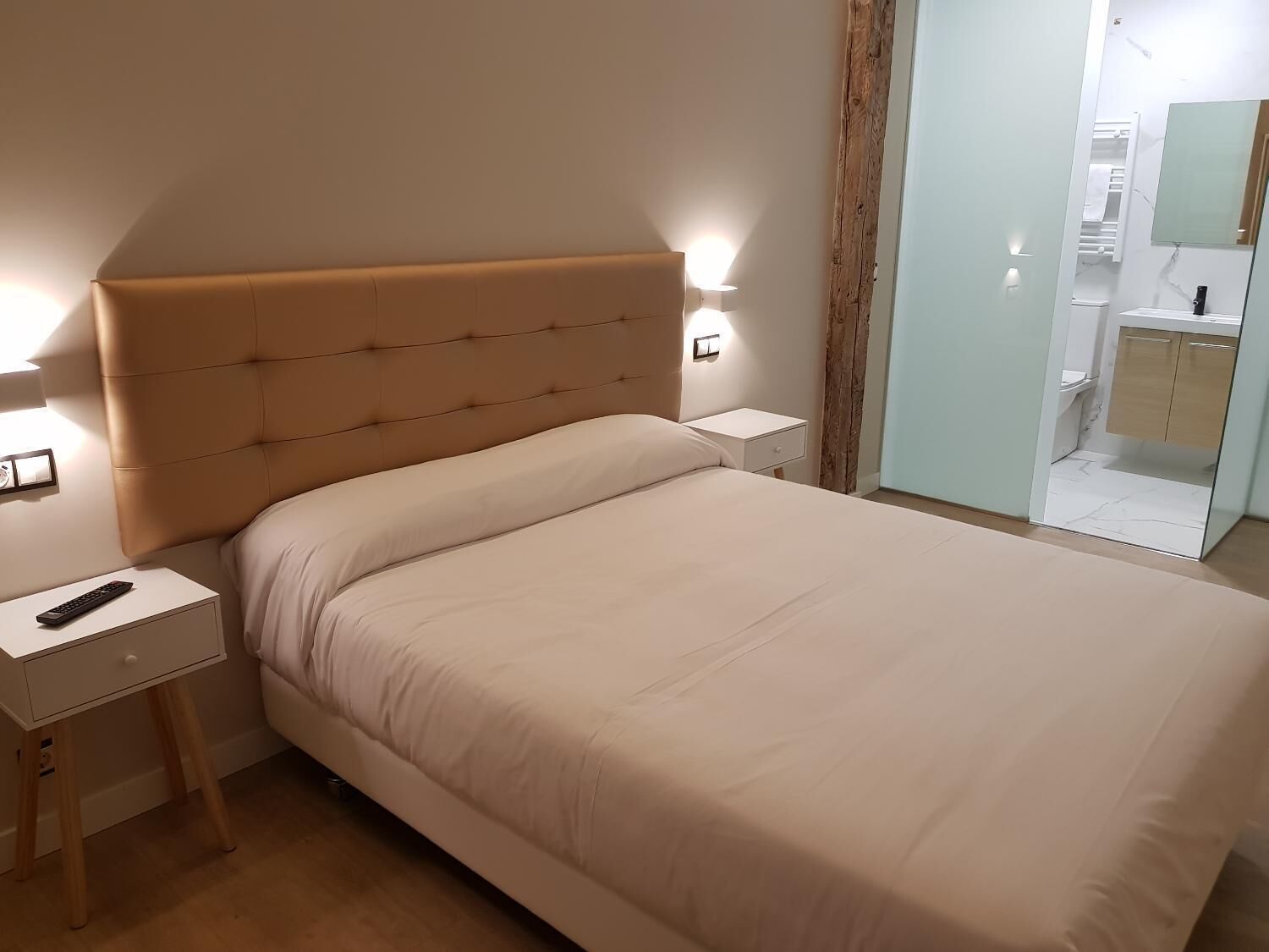A neatly made bed with a headboard, a bedside table with a remote control, and a wall-mounted light fixture.