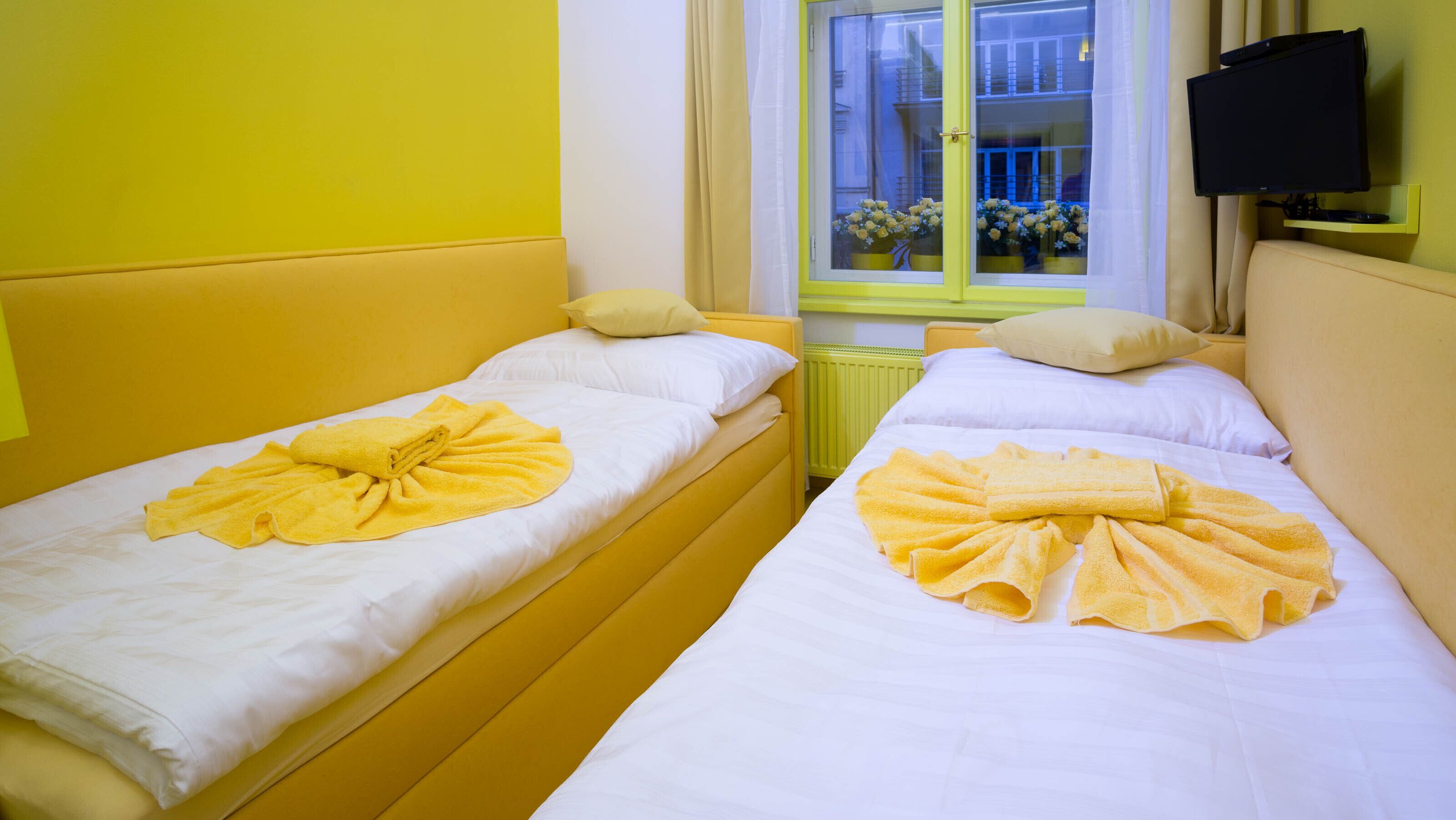 Two single beds with yellow towels, a window with flowers, and a TV on the wall.