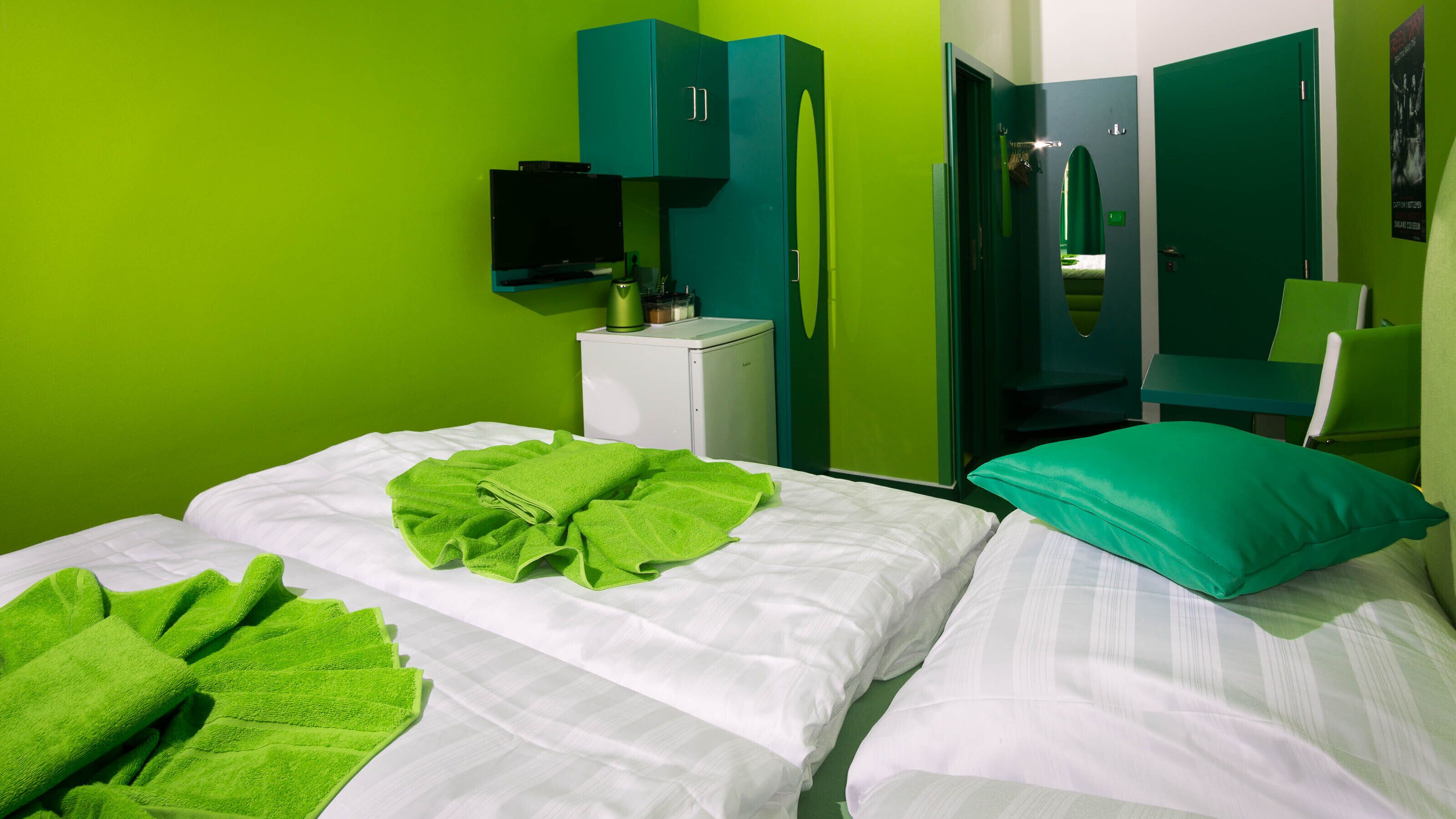 Une chambre d’hôtel avec deux lits, des serviettes vertes, une petite table de chevet blanche avec une lampe et un miroir.