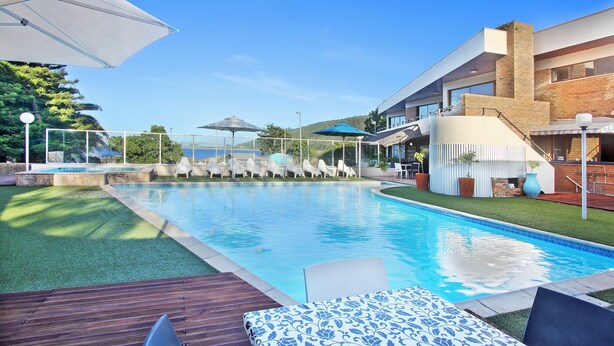A swimming pool with lounge chairs and umbrellas, surrounded by a modern building with a brick facade and a wooden deck. - Baywater Village (Sedgefield)