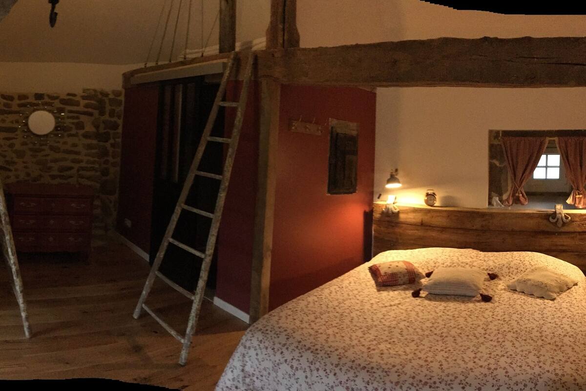 Une chambre à coucher rustique avec une échelle en bois, un lit recouvert d’une literie à motifs floraux, un mur en pierre et une tête de lit en bois.