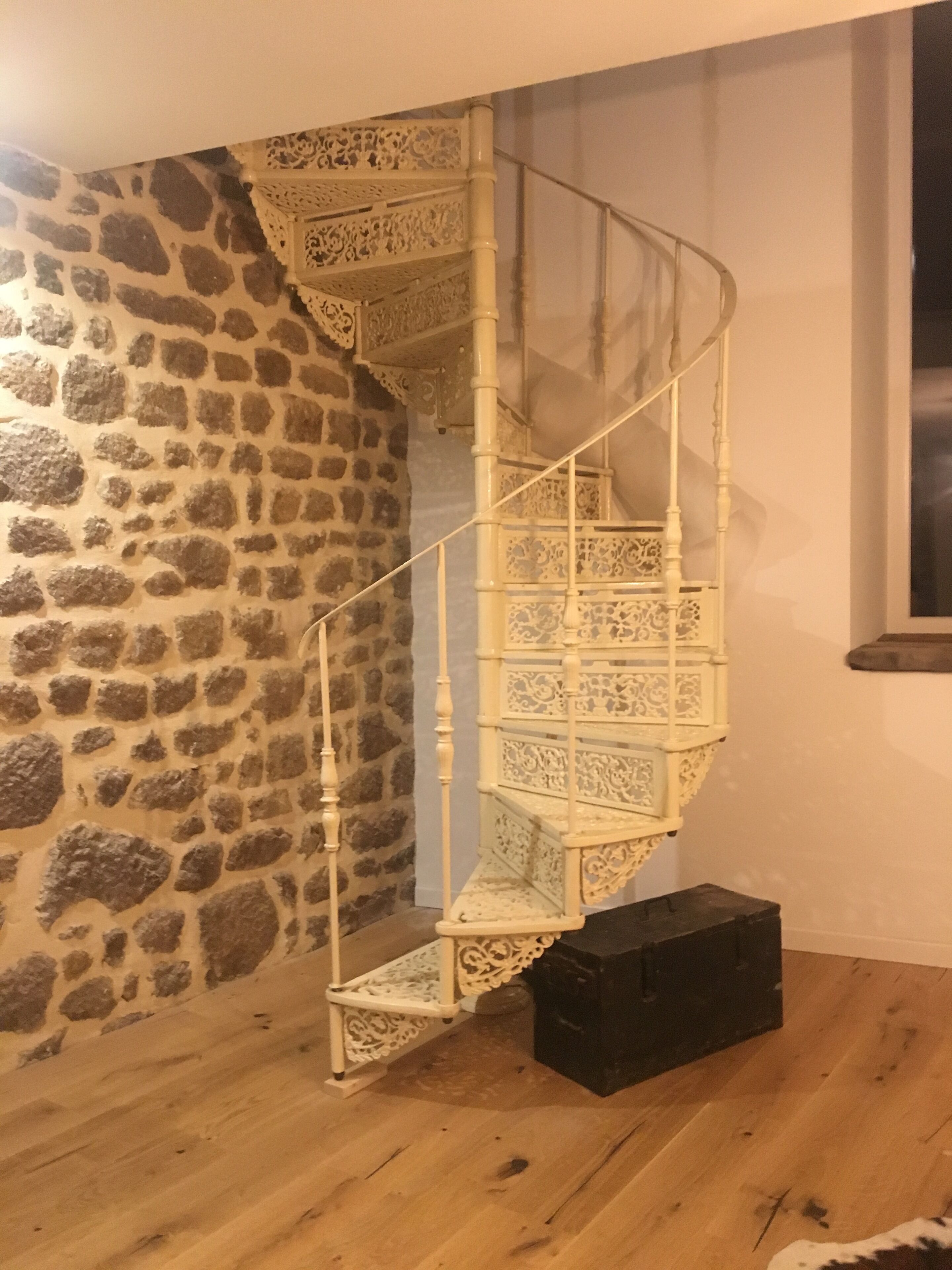 Un escalier en colimaçon aux rampes blanches finement ouvragées, adossé à un mur de pierre, mène à une sorte de boîte de couleur sombre posée sur un plancher en bois.