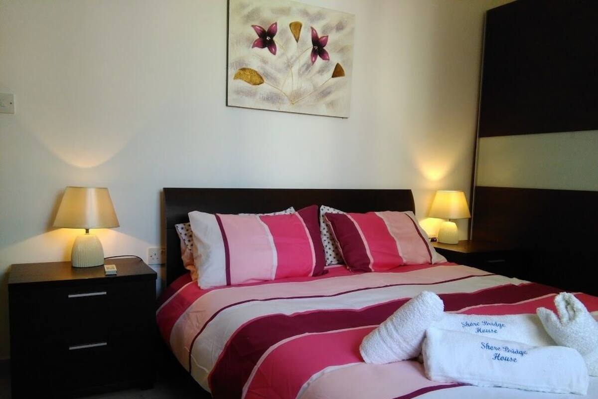 A neatly made bed with striped bedding and a white towel with text, two bedside lamps, and a wall art piece above the bed.