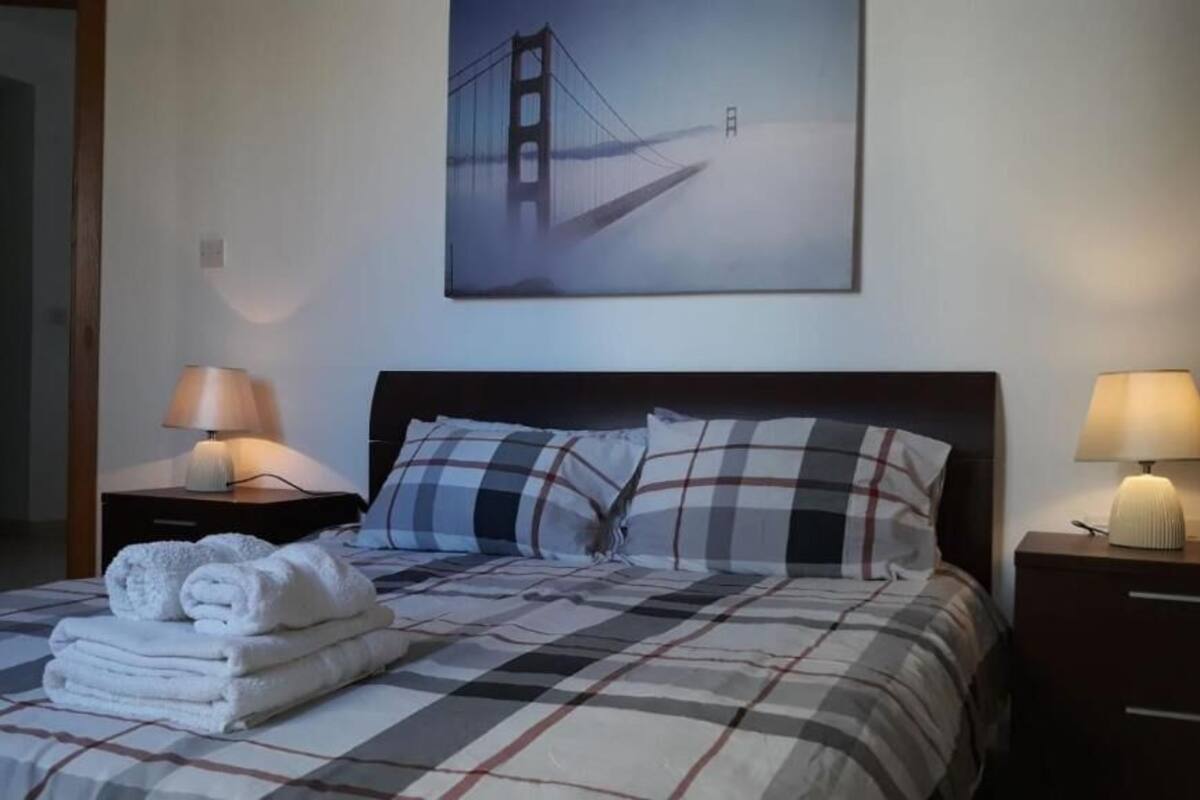 A neatly made bed with a checkered bedspread, two pillows, and a folded towel on the side. There is a framed picture of a bridge on the wall above the bed.