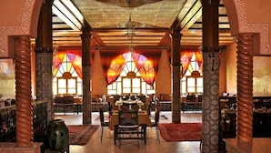 Un comedor amplio con varias mesas y sillas, ventanas grandes con cortinas estampadas y columnas decorativas.
