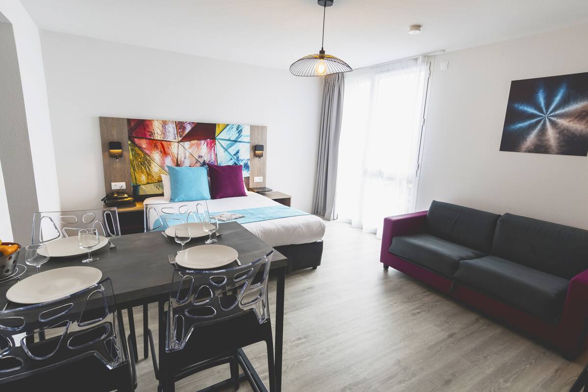 A modern hotel room with a dining area, a bed with colorful artwork, a sofa, and a large window.