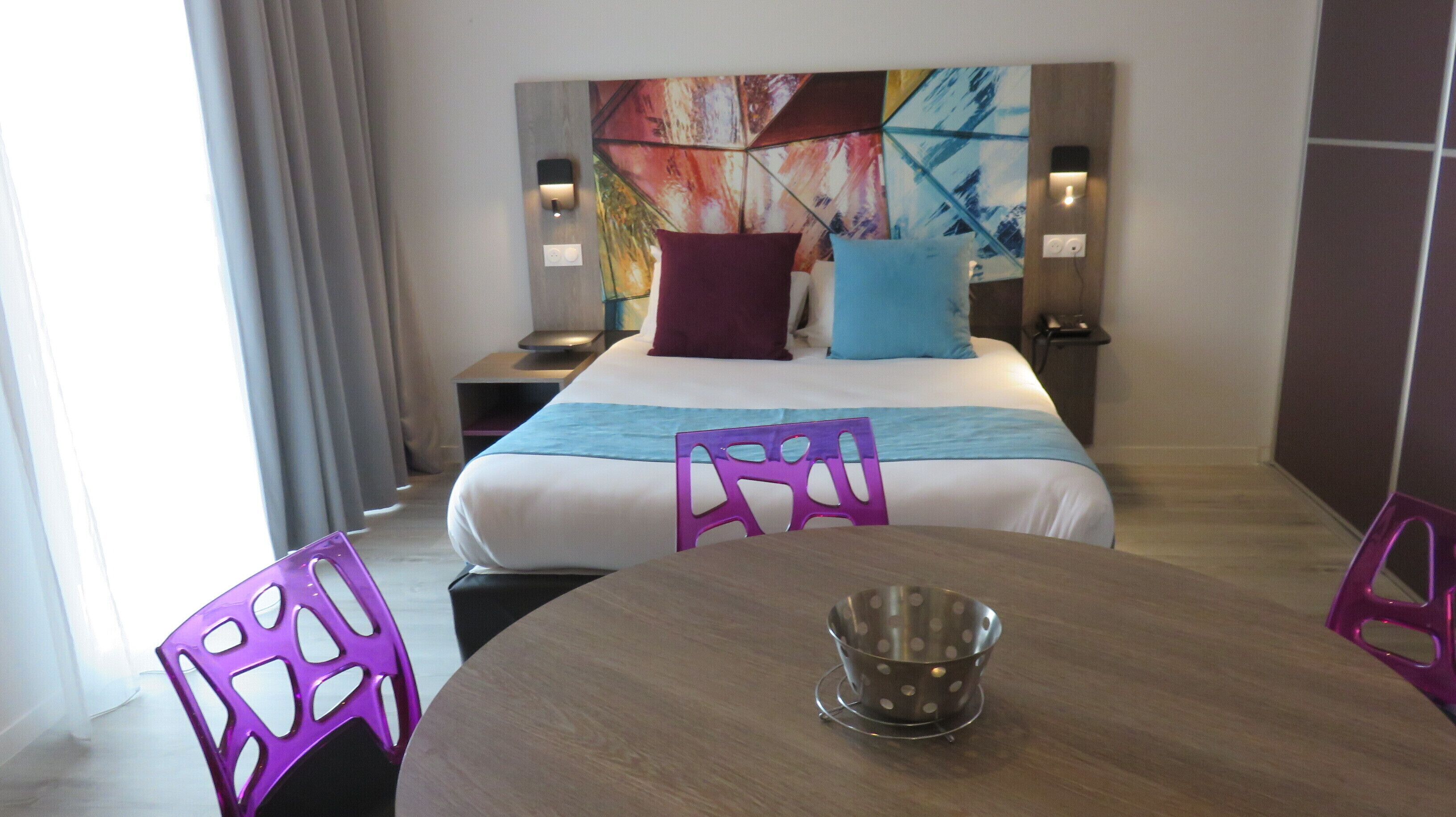 Una habitación de hotel con una cama, una mesa y sillas moradas. Hay un cuadro en la pared detrás de la cama.