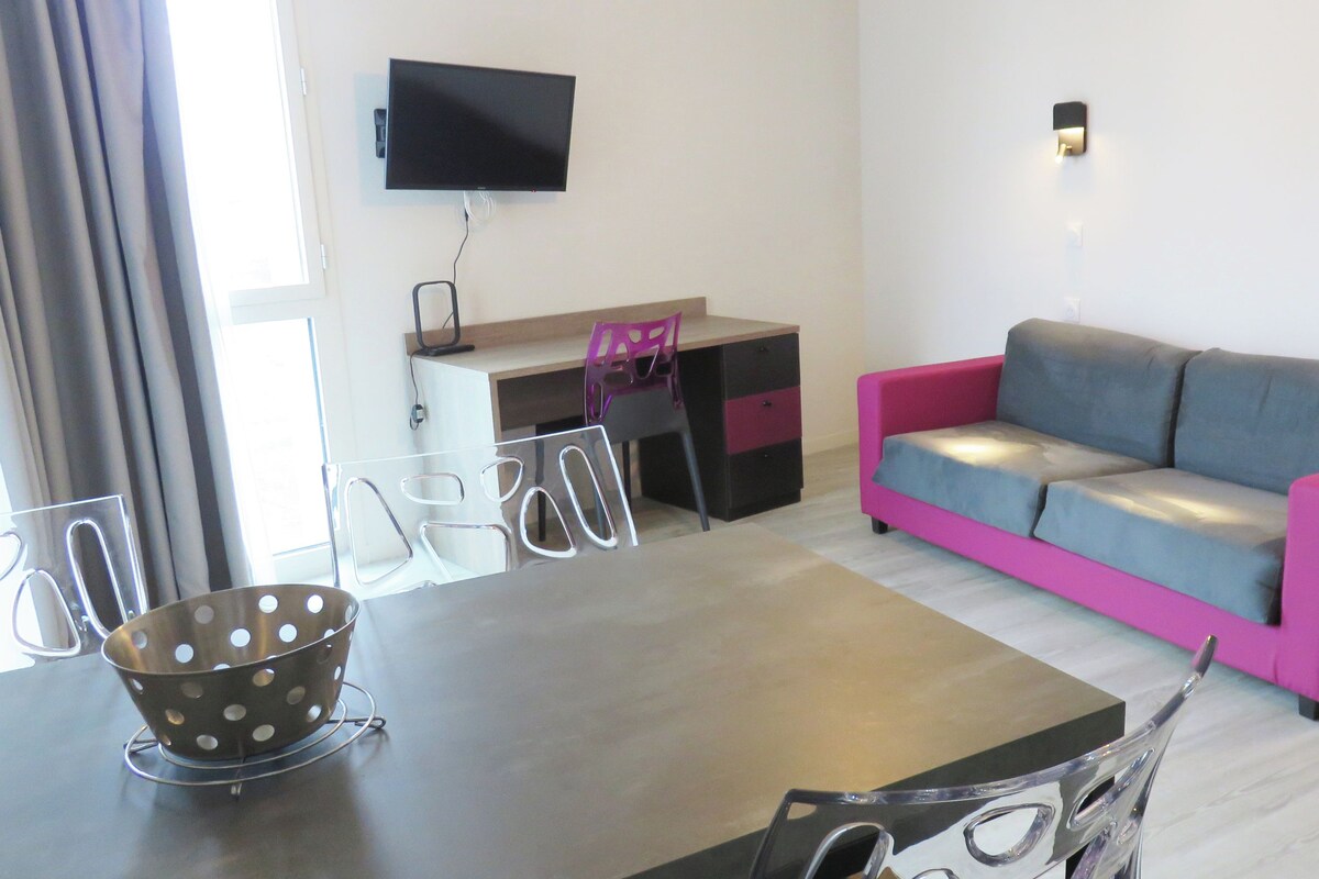 A modern living room with a flat-screen TV mounted on the wall, a pink and grey sofa, a wooden desk with a chair, and a dining table with chairs.