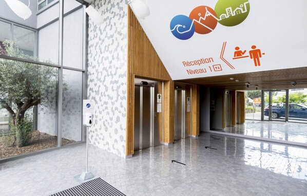 A reception area with a modern design, including a reception desk, a wall-mounted sign with icons, and a clear view of the outdoors through large windows. - Appart-Hôtel Mer & Golf City Bordeaux Lac (Bordeaux)