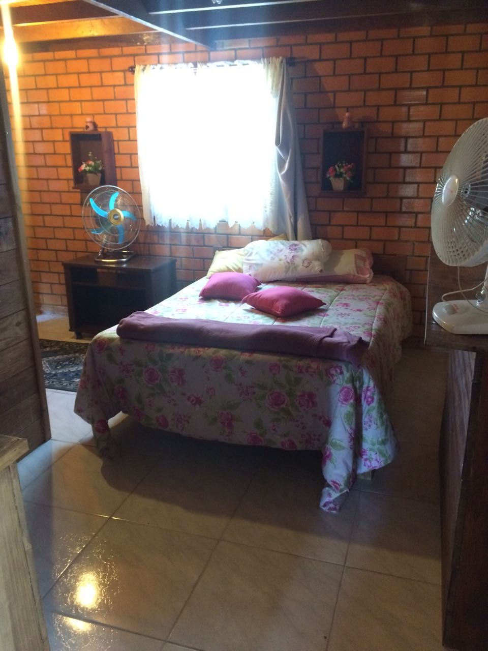 A bedroom with a brick wall, a bed with floral bedding, a standing fan, and a window with curtains.