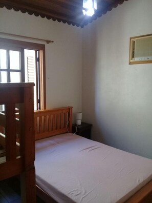 A small, single-bed room with a wooden headboard, a bedside table with a lamp, and a window with blinds. - Lagoa Country Club (Cidreira)