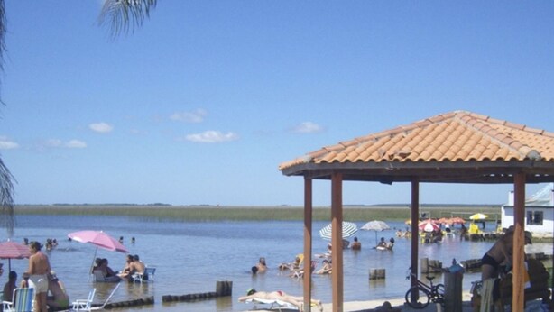 A beach with a gazebo, palm trees, and people swimming and relaxing. - Lagoa Country Club (Cidreira)