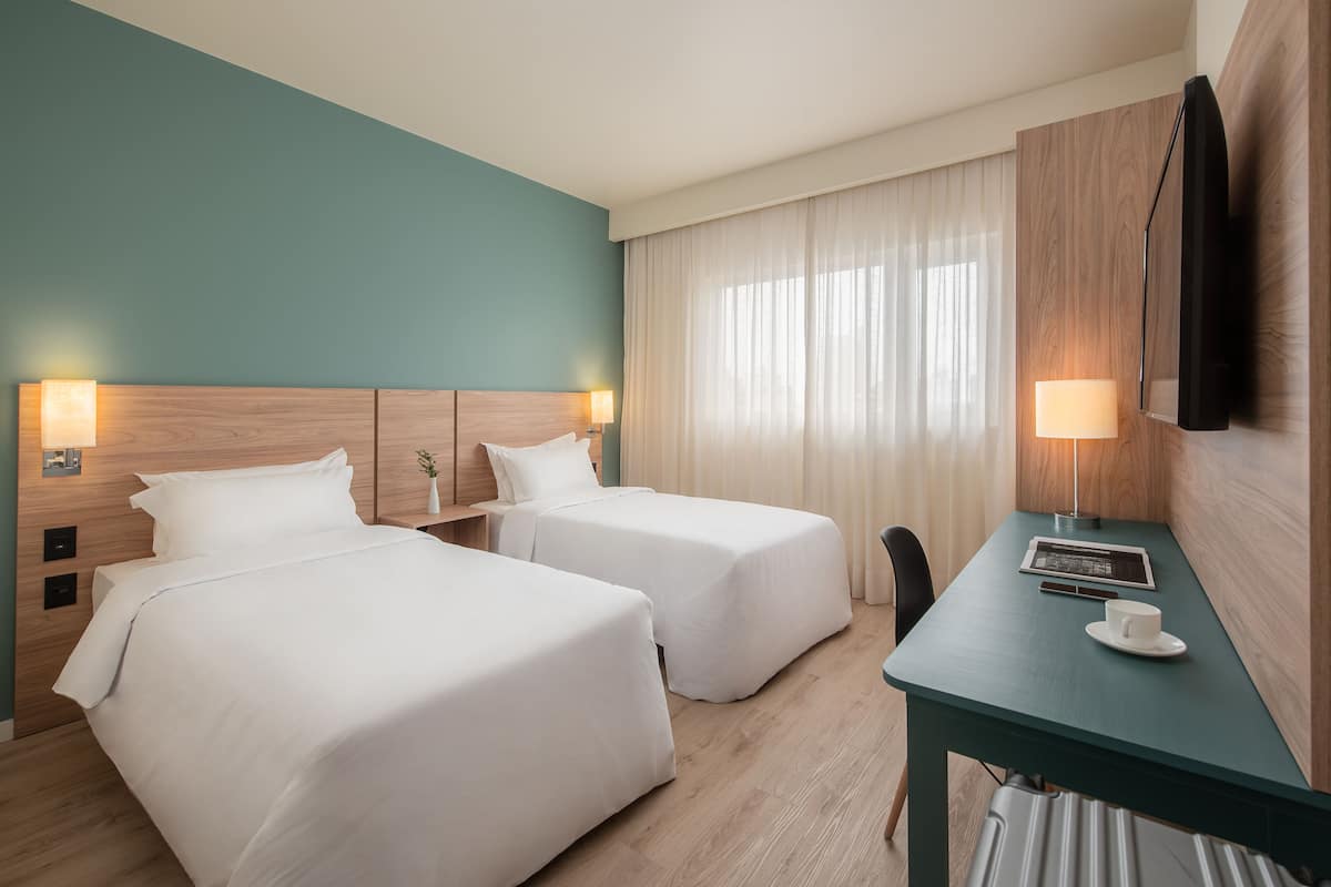 A hotel room with two beds, a desk, a TV, and a window with curtains.