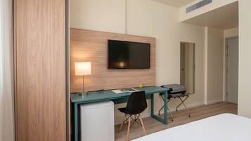 A hotel room with a bed, a desk with a computer, a TV, a lamp, and a small fridge.