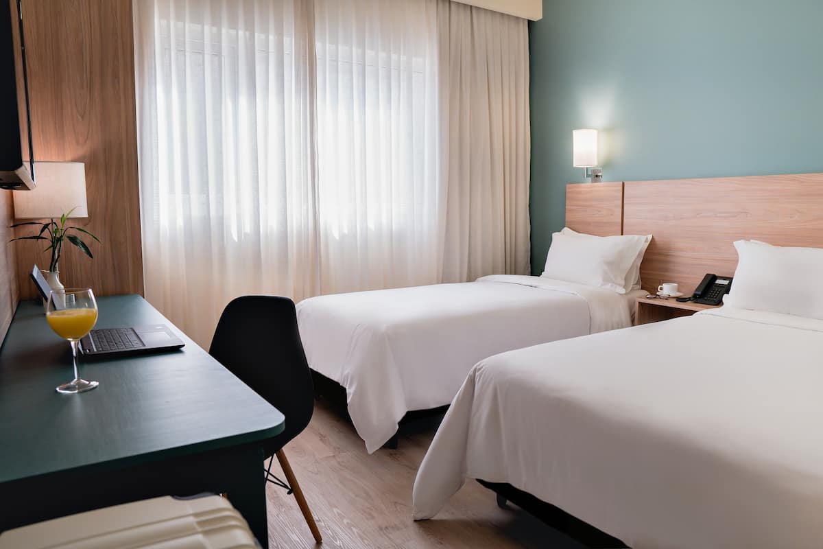 A hotel room with two beds, a desk with a laptop and a glass of orange juice, a bedside table with a lamp, and a plant.
