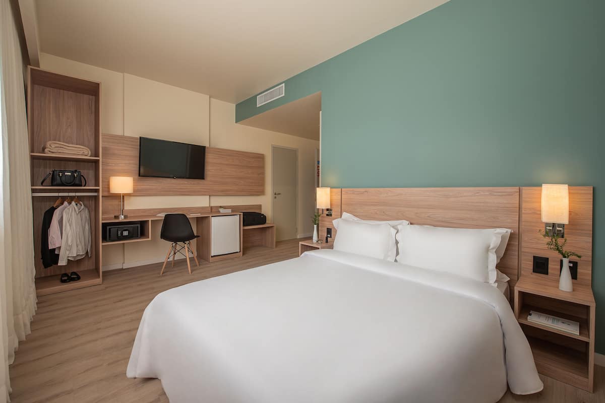 A modern hotel room with a large bed, a desk with a chair, a TV, and a wardrobe.
