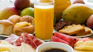 Una mesa de desayuno con diversos alimentos, como pan, queso, embutidos, frutas y un vaso de zumo de naranja.