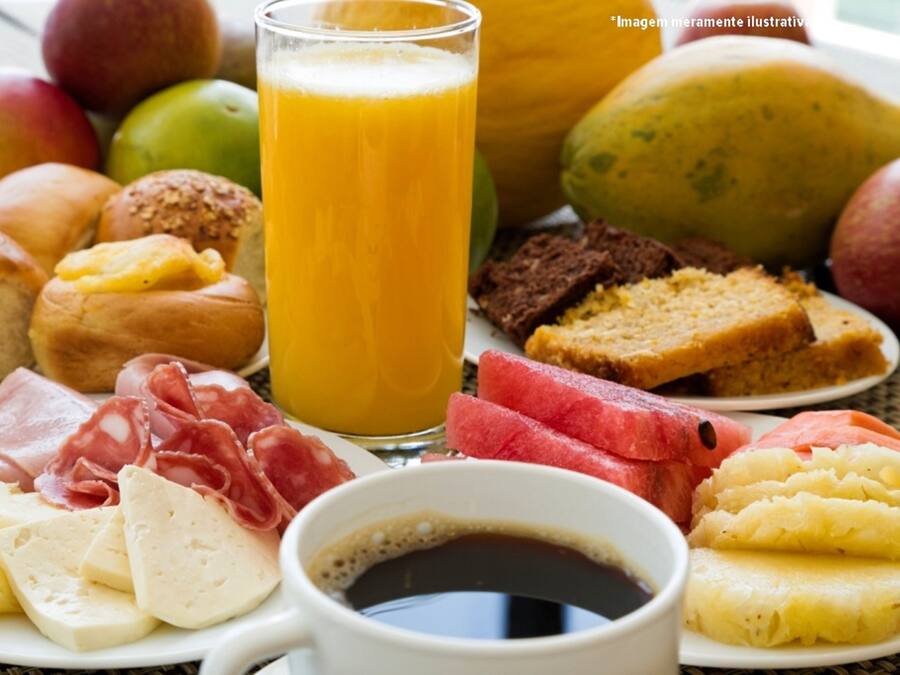 Uma mesa de café da manhã com diversos alimentos, incluindo pão, queijo, carnes, frutas e um copo de suco de laranja.