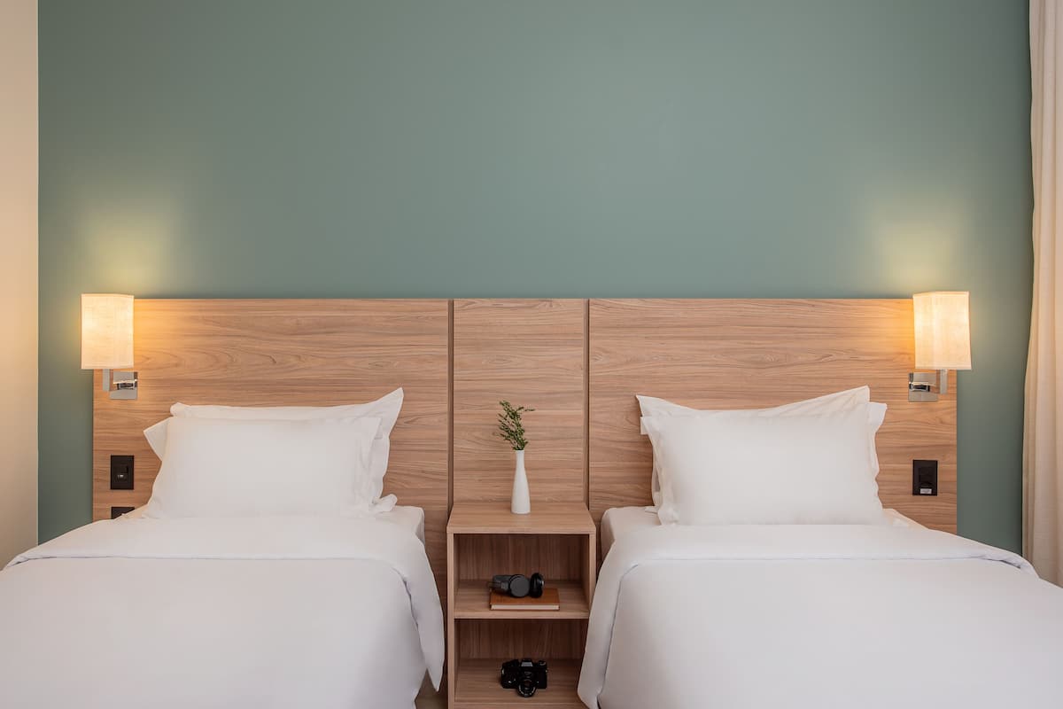 A double room with two beds, wooden headboard, bedside lamps, a small shelf with a vase and a camera, and a green wall.