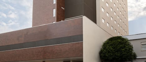 A modern building with a mix of brick and concrete facades, featuring a large entrance and a small green area.