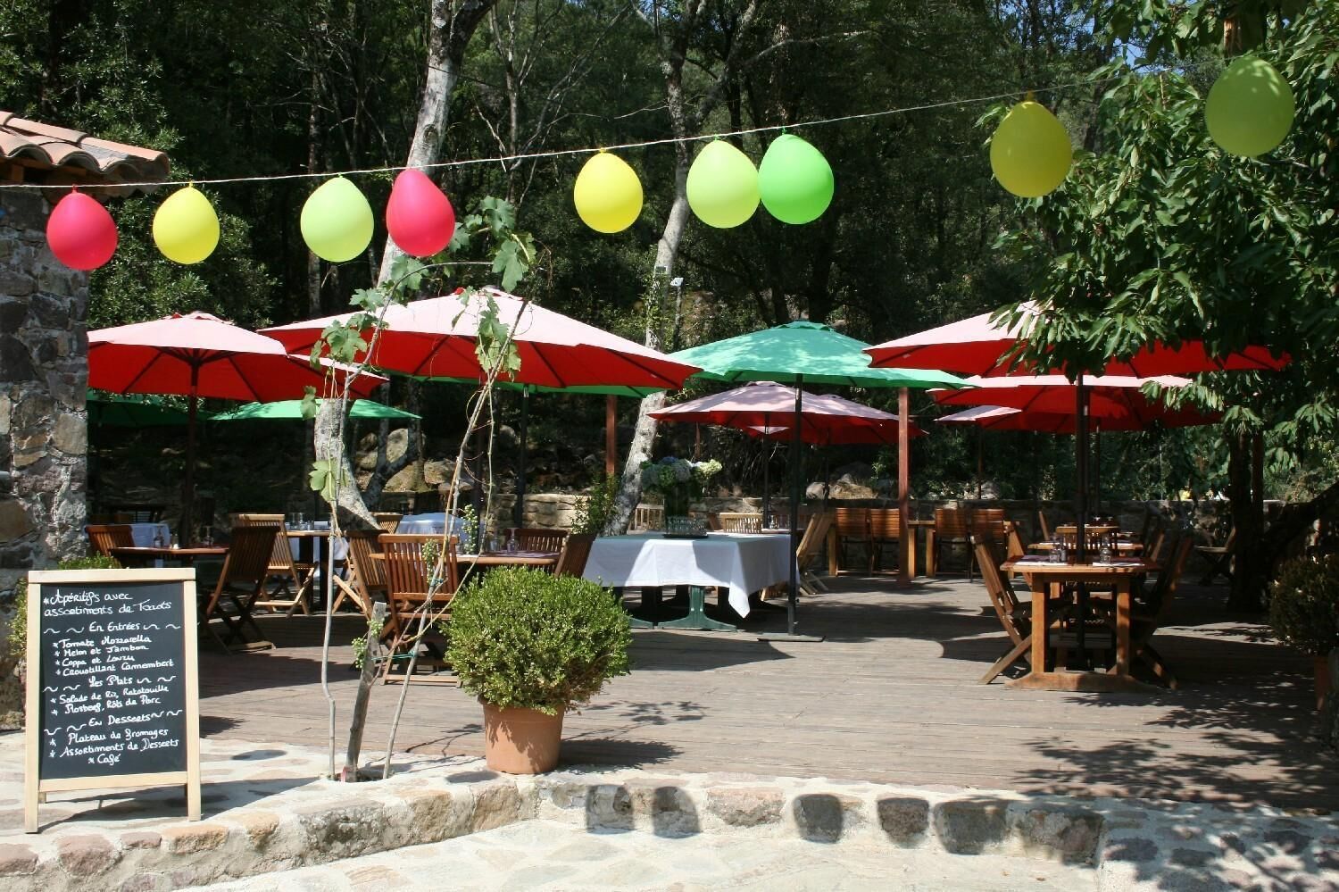 Un área de comedor al aire libre con mesas y sillas bajo sombrillas, rodeada de árboles.
