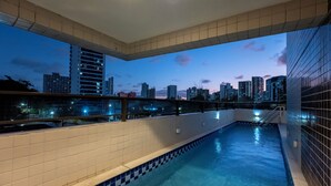 Una piscina con borde de azulejos, barandilla de vidrio y vista a un horizonte urbano al atardecer.