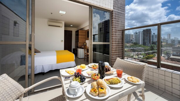 Un balcón con una mesa de comedor dispuesta con artículos para el desayuno, una cama con colcha amarilla y vistas a la ciudad a través de amplias ventanas.
