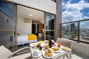 A balcony with a dining table set with breakfast items, a bed with a yellow bedspread, and a city view through large windows. - Villa Park Hotel Recife (Recife)