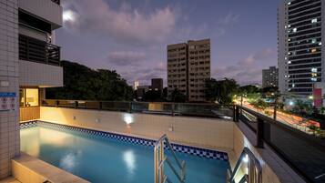 A swimming pool with a tiled deck and metal railings, surrounded by buildings and trees.