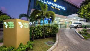 Exterior del Hotel Villapark con su letrero iluminado, entrada pavimentada y follaje circundante.