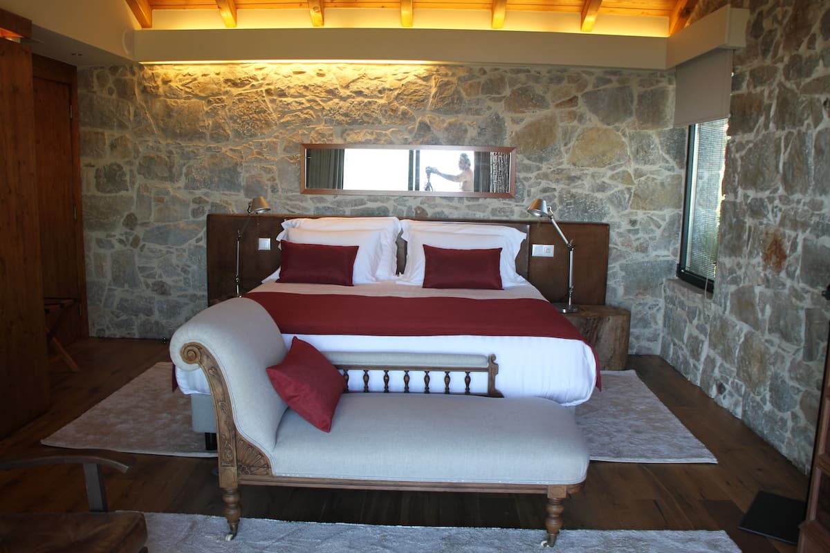 A bedroom with a stone wall, a wooden bed with red and white bedding, a wooden bench, and a nightstand with a lamp.