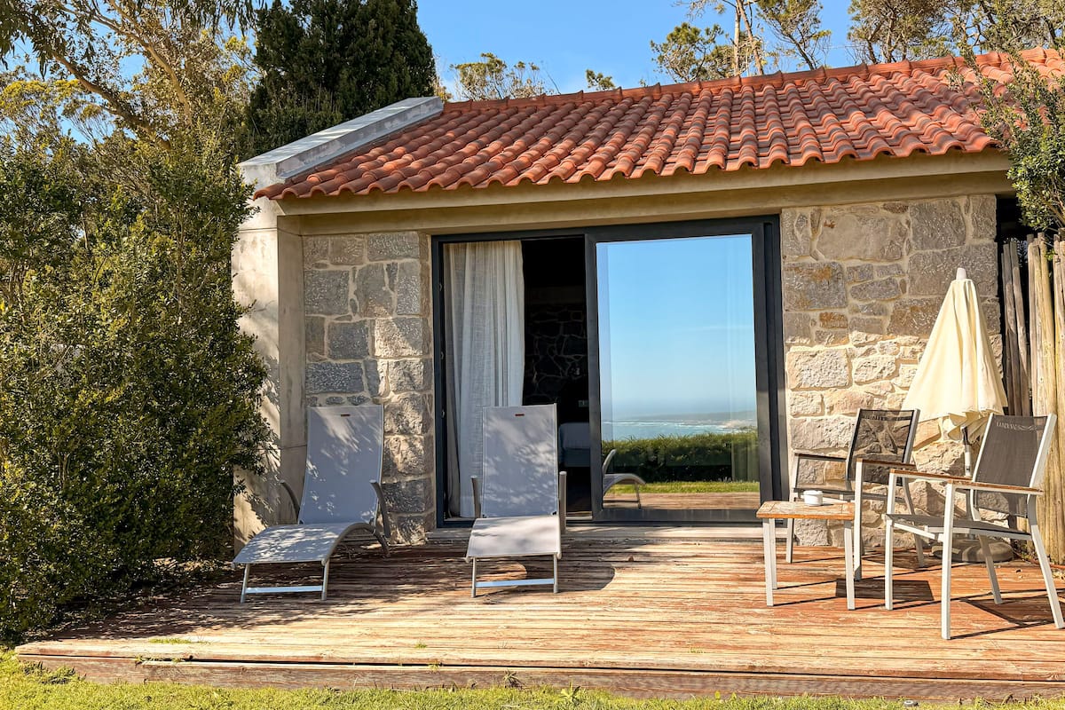 A stone building with a tiled roof, an open doorway leading to a deck, and outdoor seating with umbrellas.