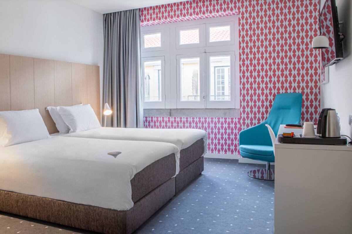 Ein Hotelzimmer mit Bett, Nachttisch, Lampe, Stuhl und Schreibtisch mit Wasserkocher und Tasse.