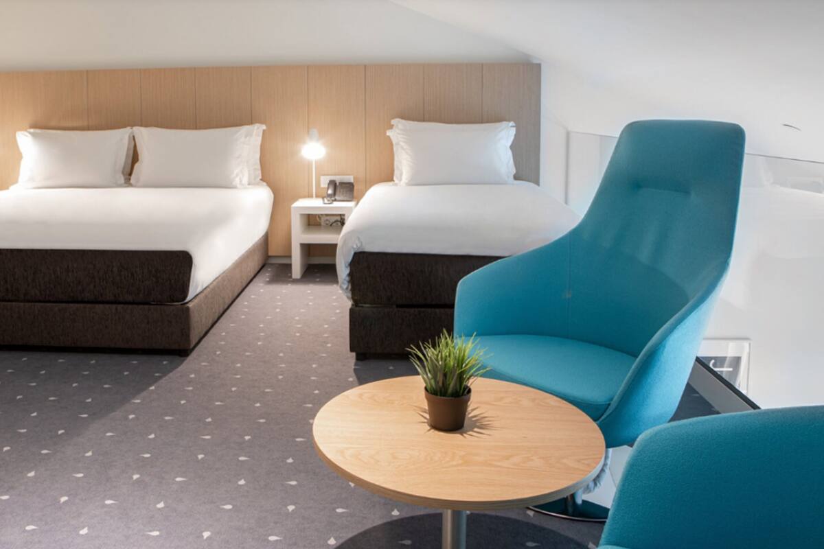 Ein Hotelzimmer mit zwei Betten, einem kleinen Nachttisch mit Lampe, einem runden Holztisch mit Pflanze und einem blauen Sessel.