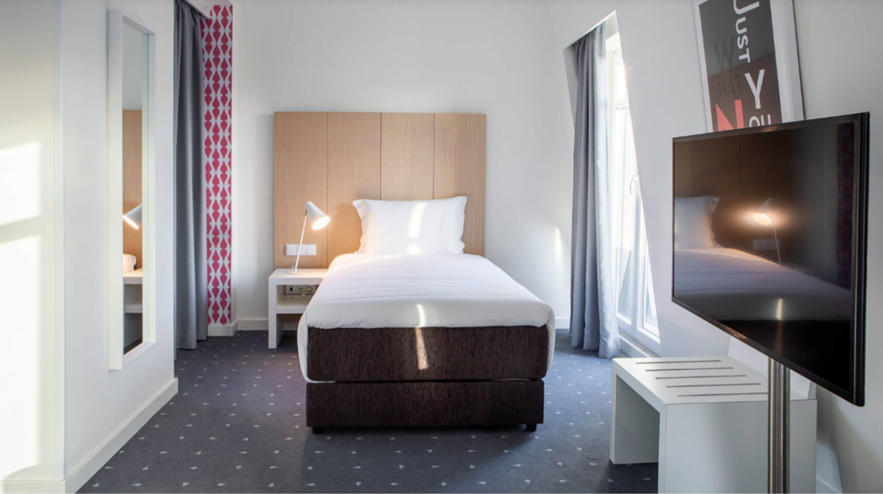 Una habitación de hotel moderna con una cama grande, una mesita de noche con lámpara, un televisor y una cortina estampada.