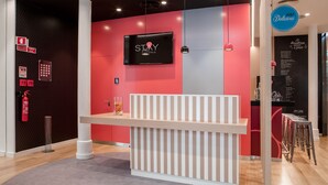 Un comptoir d’accueil doté d’un écran de télévision affichant les logos « STAY » et « HOTELS », un extincteur et un panneau indiquant un service disponible 24 heures sur 24 et 7 jours sur 7.