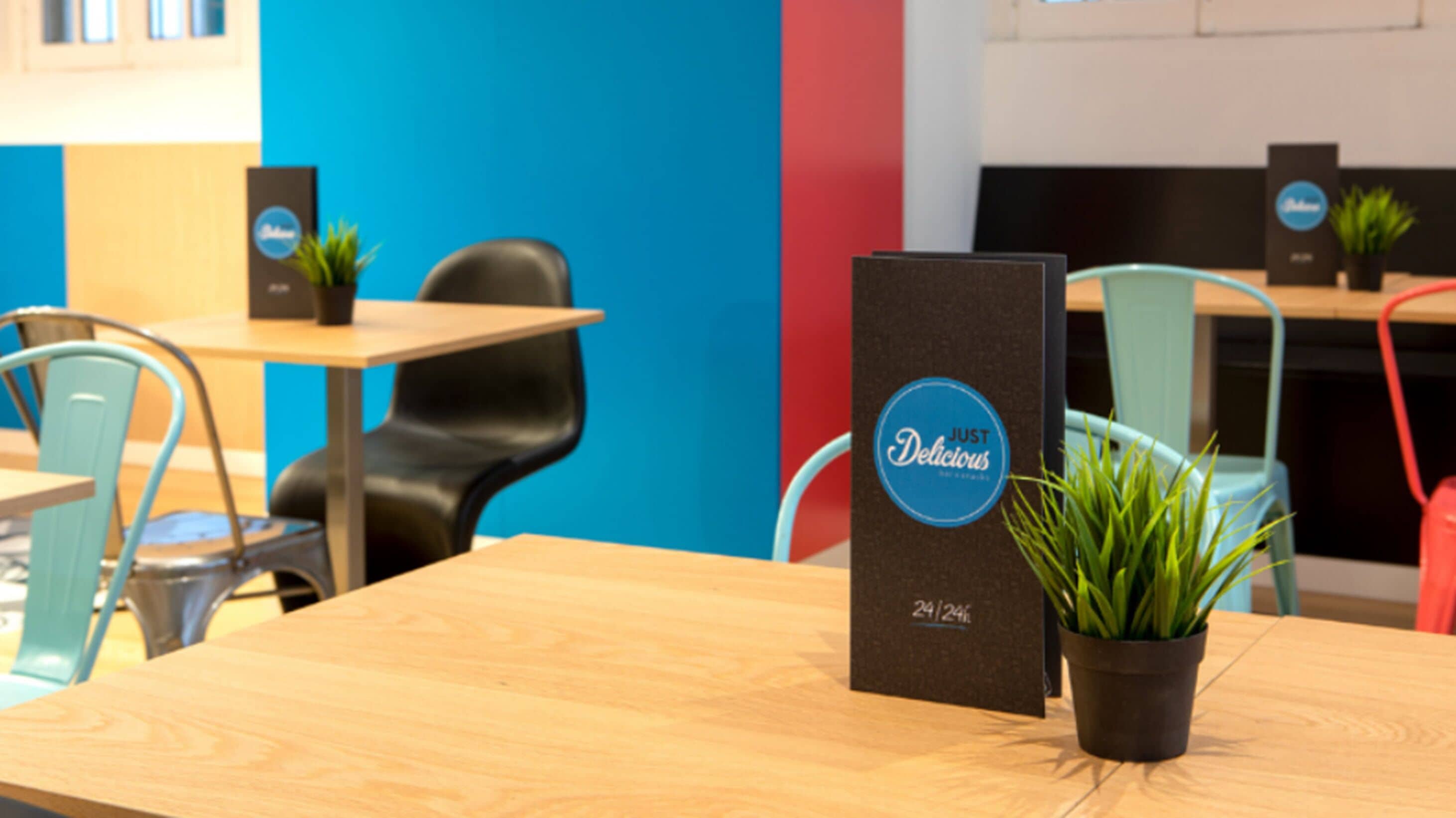 A reception desk with a sign that reads "JUST Delicious" and a logo. There are potted plants on the desk and chairs around it.