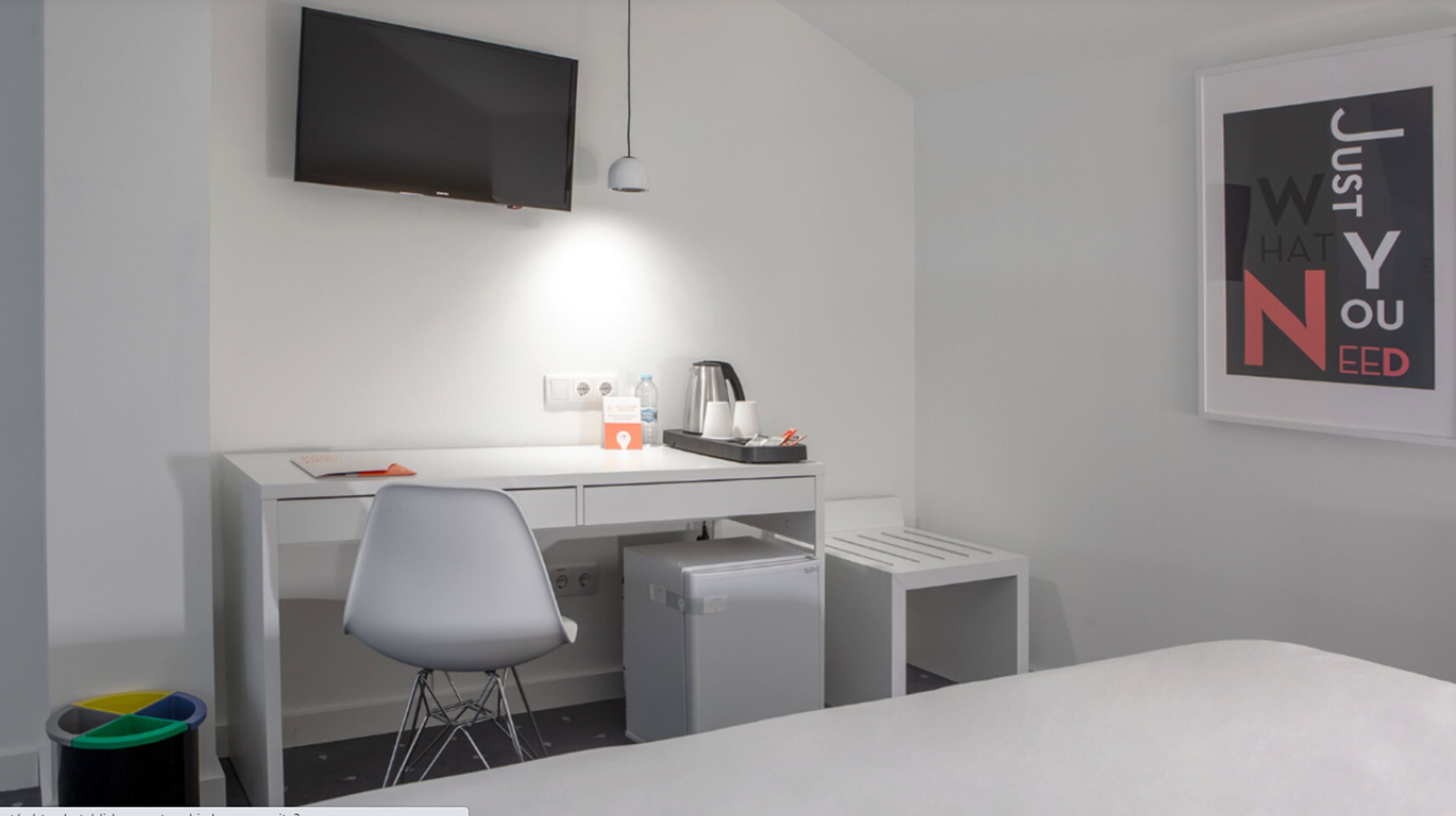 Une chambre d’hôtel moderne équipée d’un bureau blanc, d’un téléviseur à écran plat, d’un petit réfrigérateur, d’une chaise et d’une poubelle.