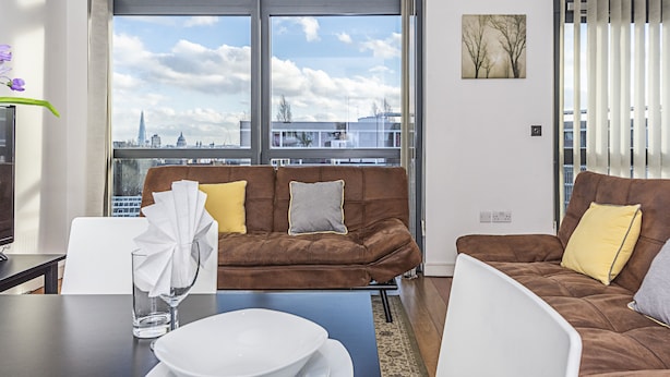Una sala moderna con un sofá marrón, una mesa de centro de cristal y vistas a un paisaje urbano a través de amplios ventanales.