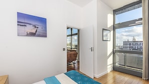 Un dormitorio con cama, suelo de madera, una puerta que da a un balcón y un ventanal con vistas a edificios y una masa de agua.