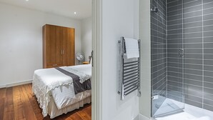 Un dormitorio con un armario de madera, una cama con colcha blanca y gris, y un baño con cabina de ducha de vidrio y un perchero.