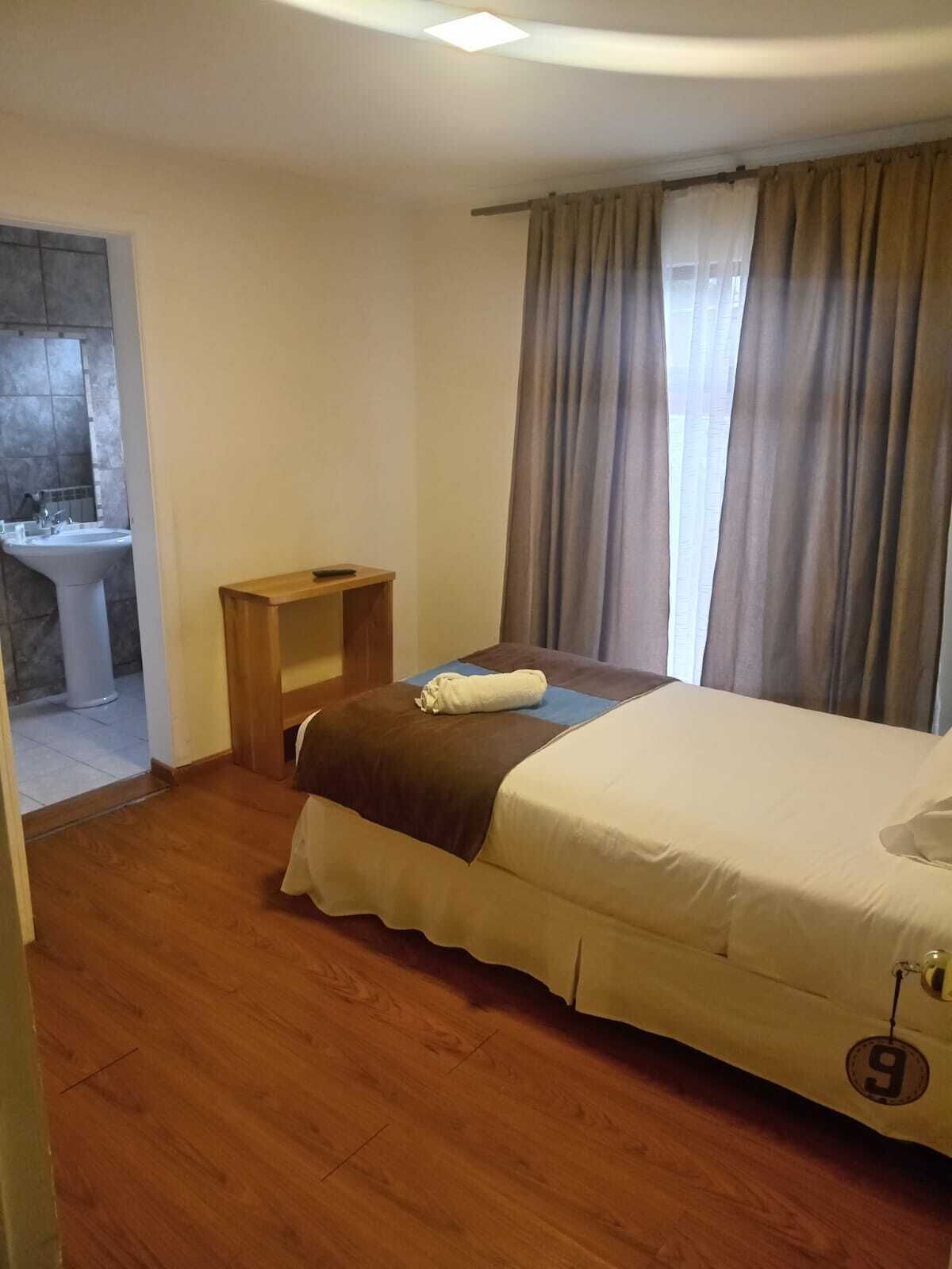 A bedroom with a bed, a wooden nightstand, a bathroom with a sink, and a window with curtains.