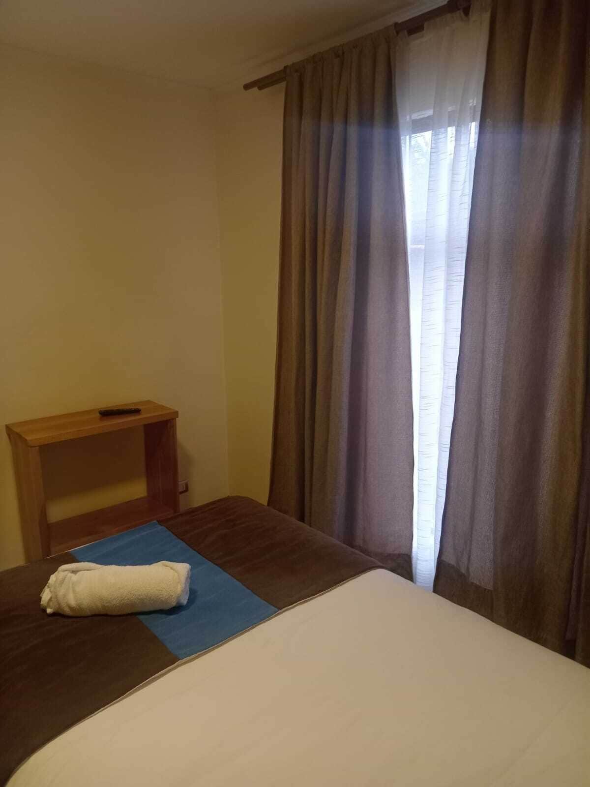 A hotel room with a bed, a wooden nightstand, and a window with curtains.