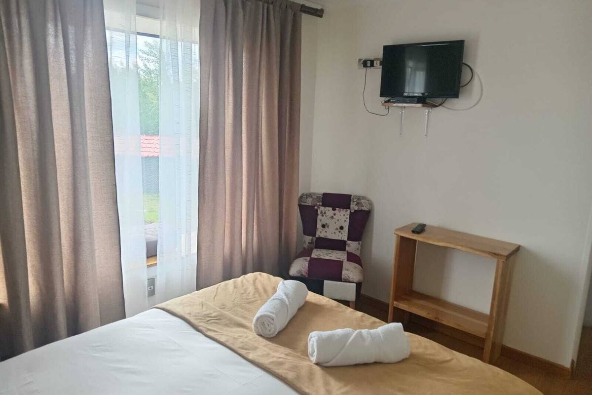 Habitación de hotel con cama, una silla, televisor montado en la pared y ventana con cortinas.