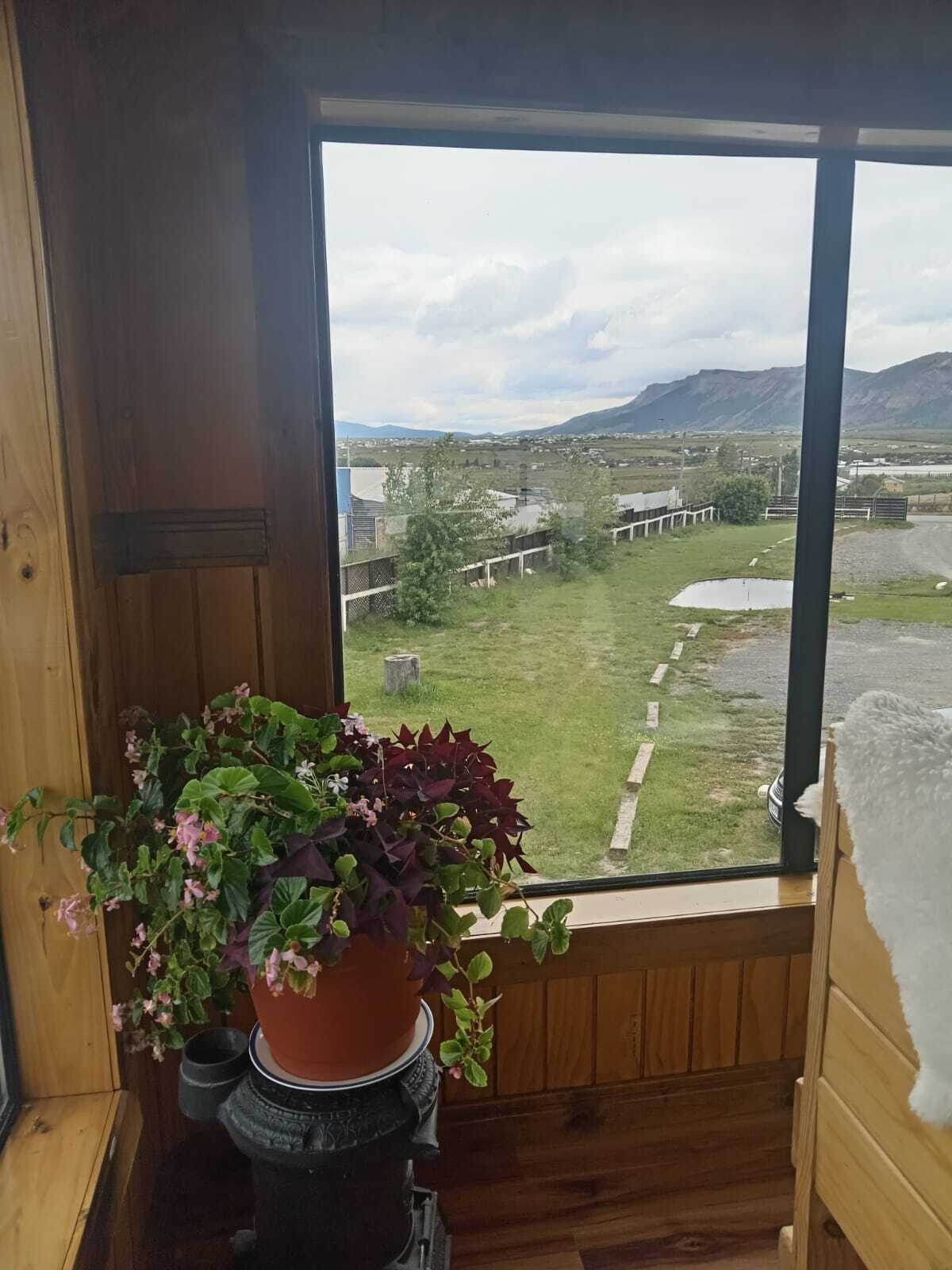 Un interior de madera con un amplio ventanal que da a un paisaje rural, una planta en maceta y una estufa pequeña negra.