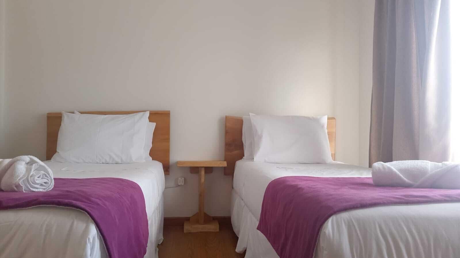 A hotel room with two beds, each with white bedding and purple throw blankets. There is a wooden nightstand between the beds.
